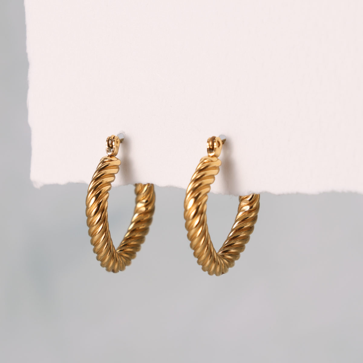 A pair of gold twisted rope hoop earrings hanging from a white display card against a soft gray background.