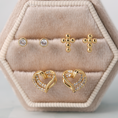 A set of gold-toned stud earrings displayed in a hexagonal beige velvet jewelry box. The set includes two small round clear-stone studs, two small cross-shaped stud earrings, and two heart-shaped stud earrings with clear stones along the edges.